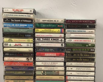 Puede incluir: Una pila de cintas de casete vintage con varios álbumes de música navideña y festiva. Los títulos incluyen "The Jingle Cats", "Christmas Favorites" y "Joy to the World". Las cintas tienen varios colores y diseños, mostrando una estética nostálgica.