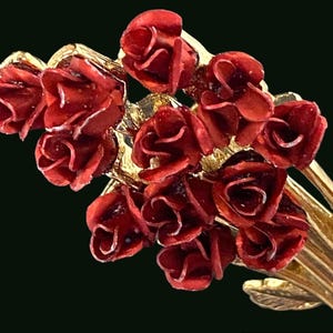 May include: A gold-toned metal brooch featuring a bouquet of red roses. The roses have detailed petals and are clustered together, with a few gold-colored leaves. The brooch is set against a dark background.