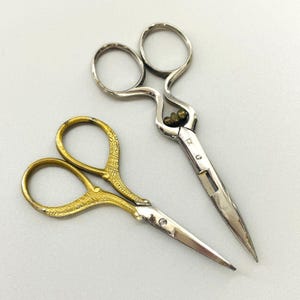 Può includere: Due paia di piccole forbici. Un paio ha manici color oro con un design ornato, e l'altro ha manici color argento. Entrambi i paia hanno lame appuntite e sono progettate per lavori di taglio di precisione.