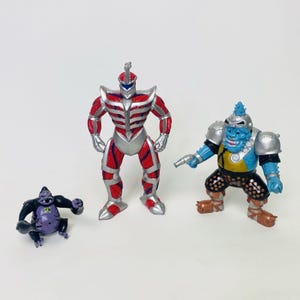 May include: Three action figures: a gorilla, a red and silver robot, and a blue monster with a gun. The gorilla is black and purple. The robot is red and silver. The blue monster has brown and black pants.