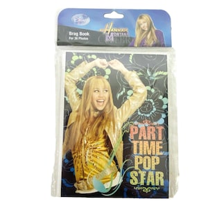 Puede incluir: Un álbum de fotos "Brag Book" de Hannah Montana para 36 fotos. La portada presenta una foto de Miley Cyrus con una chaqueta dorada y las palabras "Part Time Pop Star". El embalaje es de plástico transparente con una parte superior azul y blanca.