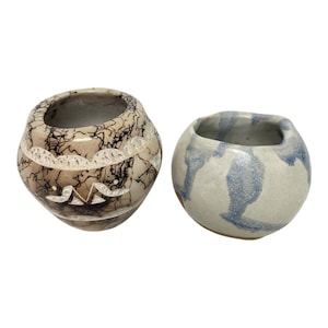 Op de afbeelding: Twee keramische kommen. Eén kom is beige met zwarte aderachtige patronen en een wit zigzagontwerp. De andere kom is lichtgrijs met blauwe aquarelachtige patronen. Beide kommen hebben een ronde vorm en open bovenkanten.