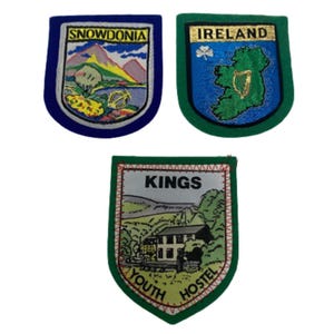 Pode incluir: Três patches bordados com temas de viagem. Um patch azul que diz "SNOWDONIA" com uma cena de montanha e lago. Outro patch verde que diz "IRELAND" com um mapa da Irlanda e um trevo. O terceiro patch diz "KINGS YOUTH HOSTEL".