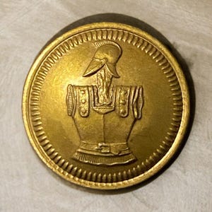 Puede incluir: Botón redondo de color oro antiguo con un diseño en relieve. El diseño presenta un busto con casco y armadura, rodeado por un borde texturizado. El botón es probablemente de metal.