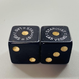 May include: Two black dice with gold-colored dots and the text "Stardust = Las Vegas" in white. The dice are square-shaped and have a vintage appearance. The dots are in the standard arrangement for dice.