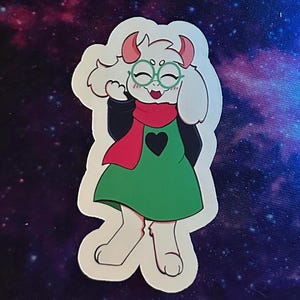 May include: A sticker depicting a cartoon character with white fur, pink horns, and green spectacles. The character is dressed in a green dress with a black heart, a red scarf, and a black top. The sticker is set against a dark, galaxy-themed backdrop.