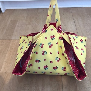 Puede incluir: Un portador de tela amarillo con estampado de fresas rojas y forro de lunares rojos. El portador tiene tres bolsillos y un asa. Está diseñado para sostener y transportar una fuente para hornear.