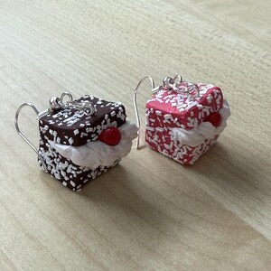 Op de afbeelding: Een paar hangende oorbellen met miniatuur taartpunten. Eén oorbel is een chocoladetaart met witte frosting en sprinkles, de andere is een roze taart met witte frosting en sprinkles.