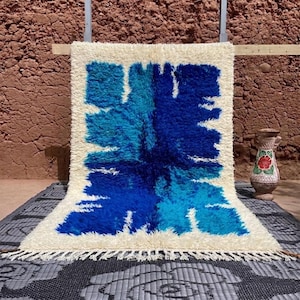 Puede incluir: Una alfombra rectangular de color crema con un diseño central azul. El diseño es abstracto, con forma de flor. La alfombra tiene flecos en el borde inferior. Se ve un jarrón decorativo con un diseño floral.
