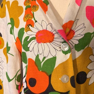May include: A long-sleeved blazer in an off-white fabric featuring a vibrant floral print with large daisies, abstract orange and yellow shapes, and green leaves. The garment has a notched lapel collar and two clear, round buttons on the front.