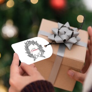K&ouml;nnte beinhalten: Ein wei&szlig;er Geschenkanh&auml;nger mit einer schwarz-wei&szlig;en Illustration eines Kranzes mit dem roten Text "Merry Christmas". Der Anh&auml;nger ist an einer braunen Geschenkbox mit einer silbernen Schleife befestigt.