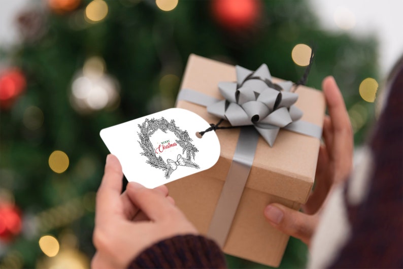 K&ouml;nnte beinhalten: Ein wei&szlig;er Geschenkanh&auml;nger mit einer schwarz-wei&szlig;en Illustration eines Kranzes mit dem roten Text "Merry Christmas". Der Anh&auml;nger ist an einer braunen Geschenkbox mit einer silbernen Schleife befestigt.