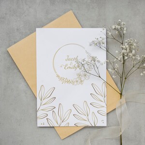 K&ouml;nnte beinhalten: Eine wei&szlig;e Hochzeitseinladung mit goldenen Lettern und floralen Designs. Die Einladung lautet "Jacob & Emily".