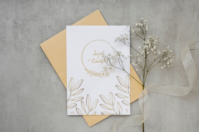 K&ouml;nnte beinhalten: Eine wei&szlig;e Hochzeitseinladung mit goldenen Lettern und floralen Designs. Die Einladung lautet "Jacob & Emily".
