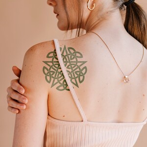 May include: A woman with a green Celtic knot tattoo on her shoulder. She is wearing a white tank top and a gold necklace with a geometric pendant.