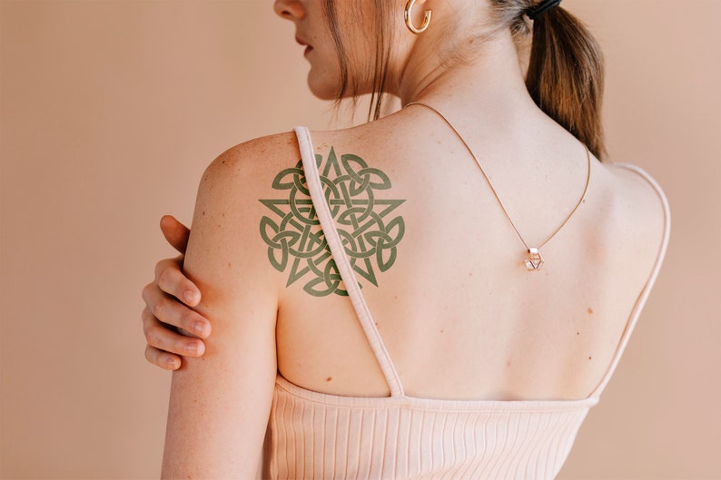 May include: A woman with a green Celtic knot tattoo on her shoulder. She is wearing a white tank top and a gold necklace with a geometric pendant.