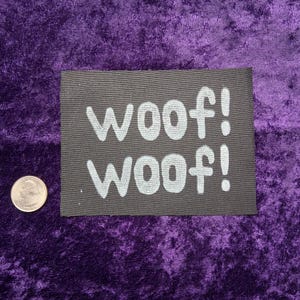 May include: A rectangular black fabric patch featuring the white text woof! woof! in a rounded sans-serif font. A silver-colored United States quarter coin is positioned to the left of the patch for scale, measuring approximately 0.955 inches in diameter.