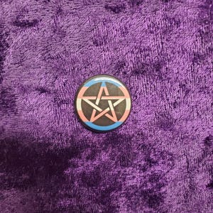May include: A round button featuring a pentagram in the centre. The pentagram is white and the background is a blue, pink and white transgender pride flag.