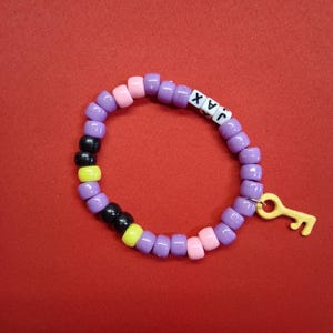 May include: A circular beaded bracelet featuring purple, pink, yellow, and black plastic pony beads. The bracelet includes white letter beads spelling the name JAX and a yellow plastic key-shaped charm attached to the side with a metal jump ring.