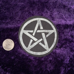 May include: A circular black fabric patch featuring an embroidered silver pentagram design. The patch is positioned next to a United States quarter coin, which has a diameter of approximately 0.955 inches, providing a clear scale for the item.