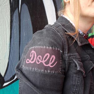 May include: A dark gray denim jacket with a pink patch that says "Doll" in cursive. The patch is stitched onto the jacket's sleeve. The jacket has pockets and a collar.
