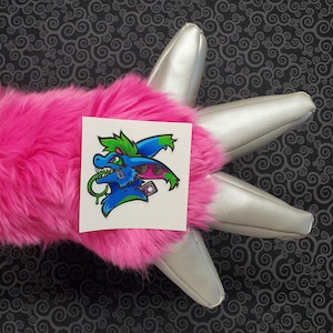 May include: A sticker featuring a blue and green cartoon creature with a pink and black design. The creature has a green tongue sticking out and a black eye. The sticker is on a pink furry surface.
