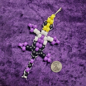 Może przedstawiać: Breloczek z jaszczurką zrobiony z koralików w kolorze fioletowym, czarnym, białym i żółtym. Jaszczurka stoi prosto, a jej ogon jest zwinięty.