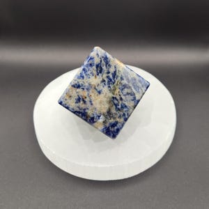 May include: A blue and white square-shaped crystal pyramid on a white selenite charging plate. The crystal has a marbled pattern of blue and white.