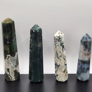 May include: Four polished gemstone obelisks in shades of green, white, and blue. The obelisks have a faceted design and stand upright on a dark surface against a white background. Each obelisk has a pointed top.