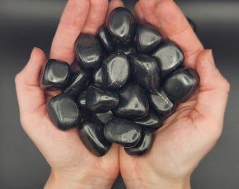 May include: A collection of smooth, black tumbled stones, likely obsidian, held in cupped hands. The stones are various sizes and shapes, with a glossy finish.