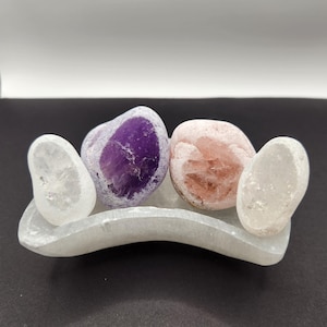 Puede incluir: Tres piedras pulidas, una morada, una rosa y una blanca, se muestran en una placa de carga de selenita blanca.
