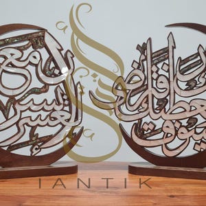 Peut inclure: Deux pièces décoratives en bois avec calligraphie arabe, chacune dans un croissant de lune. La calligraphie est d'une couleur plus claire, contrastant avec le bois brun foncé. La base est rectangulaire. Le mot "IANTIK" est en bas.