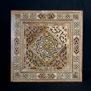 Peut inclure: Boîte décorative carrée en bois avec des motifs géométriques complexes et des détails incrustés. La boîte présente un motif central en étoile et le mot "ANTIK" au centre. La boîte est faite de différentes teintes de bois et incrustée de petits morceaux de différentes couleurs.