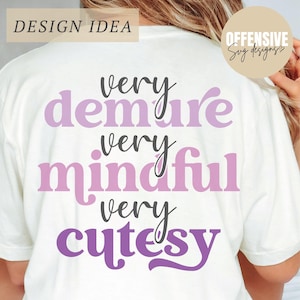 May include: Cream-colored t-shirt with the words "very demure, very mindful, very cutesy" in shades of purple and black. The shirt has the text "DESIGN IDEA" in the upper left corner and a logo in the upper right corner.