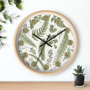 Könnte beinhalten: Eine runde Uhr mit Holzrahmen, weißem Zifferblatt und grünem Farn- und Blattmotiv. Das Zifferblatt der Uhr hat schwarze Zeiger und schwarze römische Ziffern als Stundenmarkierungen.