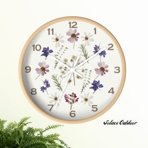 May include: Round wall clock with a light wood frame. The clock face is white, adorned with dried flowers in shades of purple, white, and green. Black numbers mark the hours. The clock hands are white. The text "Solace Outdoor" is at the bottom right.