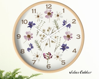 May include: Round wall clock with a light wood frame. The clock face is white, adorned with dried flowers in shades of purple, white, and green. Black numbers mark the hours. The clock hands are white. The text "Solace Outdoor" is at the bottom right.