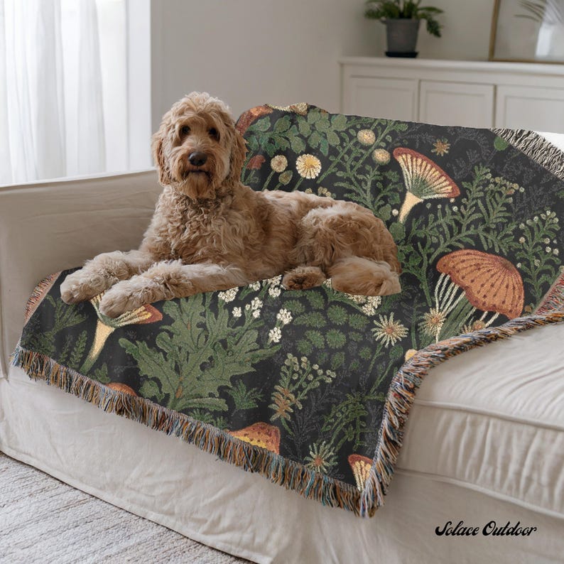 K&ouml;nnte beinhalten: Eine dunkel gef&auml;rbte Decke mit einem botanischen Design mit Pilzen und Bl&auml;ttern. Die Decke hat einen Fransenrand und ist &uuml;ber ein wei&szlig;es Sofa drapiert. Ein goldfarbener Hund liegt auf der Decke.