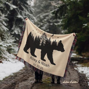 May include: Beige blanket with a black bear silhouette, forest, and moon design. The blanket has a fringed edge with red and black accents. Text on the blanket reads "Riley & Noah" and "10.10.25".