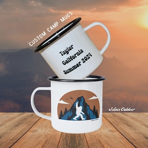 May include: Two white camp mugs with black rims and handles are stacked. One mug has text that reads "Taylor California Summer 2024." The other mug features a Bigfoot graphic with a mountain and tree design. The words "Custom Camp Mugs" are above the top mug.