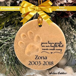 Puede incluir: Un adorno navide&ntilde;o de cer&aacute;mica beige con una huella de pata y el texto "paws leave prints on our hearts that never wash away" y "Zona 2003-2018".