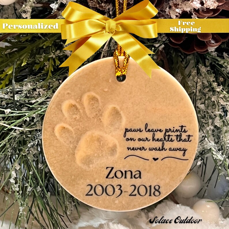 Puede incluir: Un adorno navide&ntilde;o de cer&aacute;mica beige con una huella de pata y el texto "paws leave prints on our hearts that never wash away" y "Zona 2003-2018".