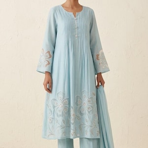 May include: A light blue long-sleeved tunic with a notched neckline and vertical pleats, featuring white floral embroidery with silver accents on the sleeves and hem, paired with matching straight-leg trousers and a coordinating light blue scarf.