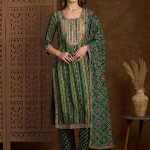 Puede incluir: Traje salwar kameez con estampado verde y blanco con una dupatta a juego. El traje presenta un escote redondo con bordados y mangas tres cuartos. Los pantalones son de un verde más oscuro con un patrón similar. El atuendo se usa con sandalias doradas.