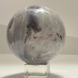 May include: A polished spherical stone featuring a marbled pattern of white, grey, and soft pink tones with dark inclusions. The smooth, rounded object rests securely on a small, clear, square acrylic stand, showcasing its natural mineral texture.