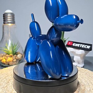 Op de afbeelding: Een blauwe ballondier-sculptuur in de vorm van een hond. Het beeld staat op een zwarte ronde basis met een reflecterend oppervlak.