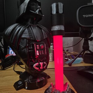 Op de afbeelding: Een zwart Darth Vader-buste met een rood lichtzwaard ernaast. Het lichtzwaard is aan en gloeit rood. De buste staat op een zwarte basis met een zilveren ontwerp. Het lichtzwaard staat op een zwarte basis met een zilveren ontwerp dat eruitziet als rotsen.