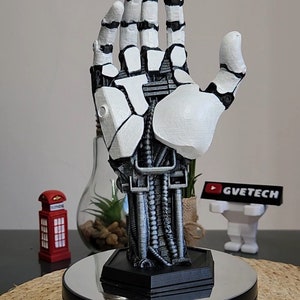 Może przedstawiać: Rzeźba robota dłoni w kolorze białym i szarym z czarną podstawą. Dłoń jest uniesiona z wyciągniętymi palcami. Podstawa ma srebrną, odblaskową powierzchnię.