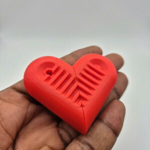 Puede incluir: Un objeto rojo brillante en forma de coraz&oacute;n con una superficie texturizada y un peque&ntilde;o agujero en la parte superior. El coraz&oacute;n est&aacute; dividido por la mitad. El objeto se sostiene en una mano sobre un fondo liso.