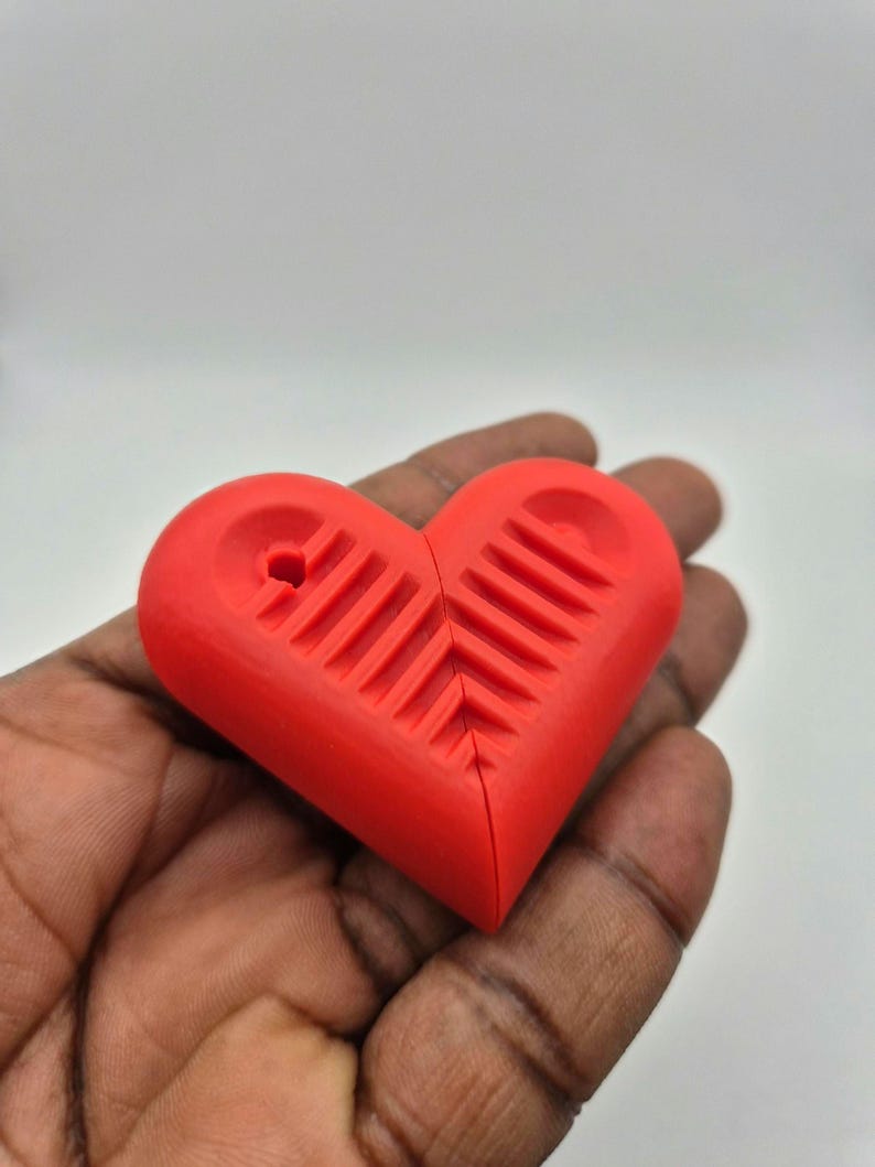 Puede incluir: Un objeto rojo brillante en forma de coraz&oacute;n con una superficie texturizada y un peque&ntilde;o agujero en la parte superior. El coraz&oacute;n est&aacute; dividido por la mitad. El objeto se sostiene en una mano sobre un fondo liso.